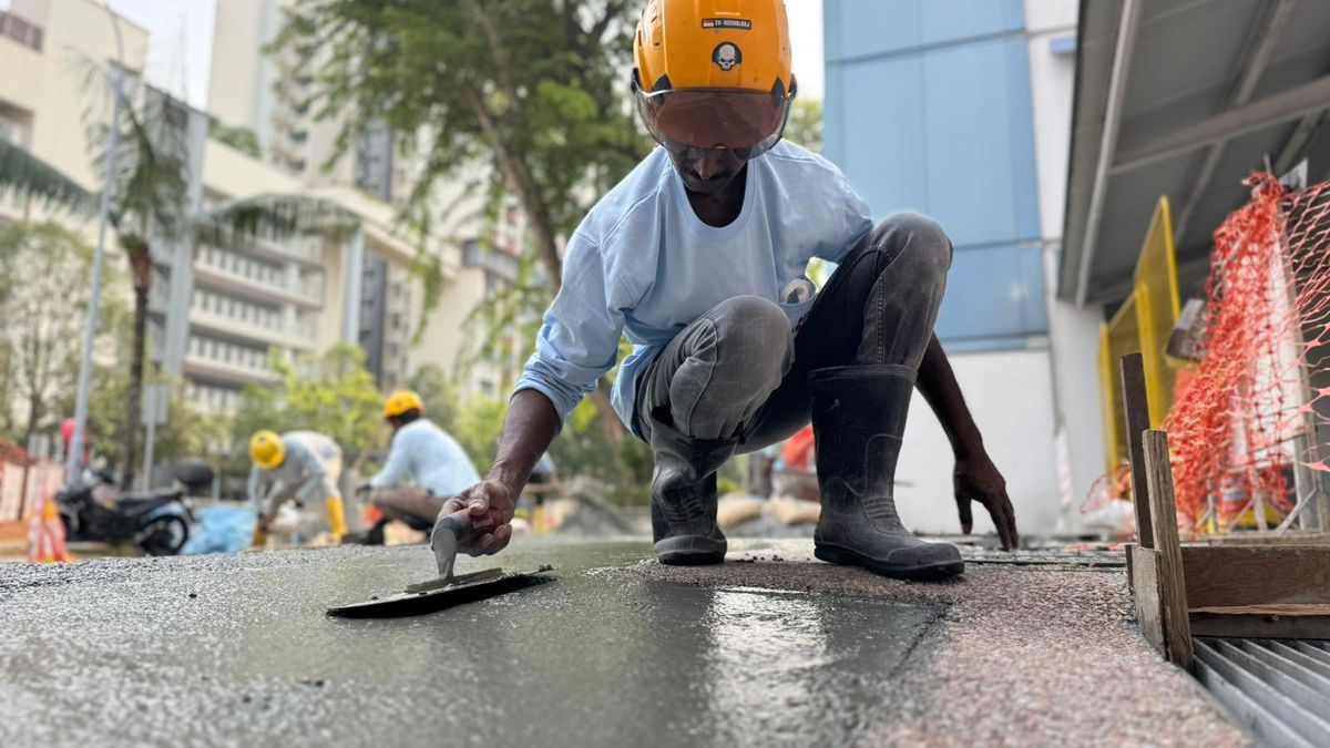 Worker in a yellow hard hat kneeling to hand-finish a fresh surface with a steel trowel, with other workers and residential blocks behind.
