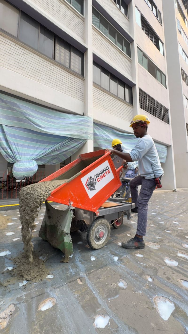 Worker tipping an orange dumper with a Rhino Crete logo panel, discharging wet mix onto a surface beside a residential block.
