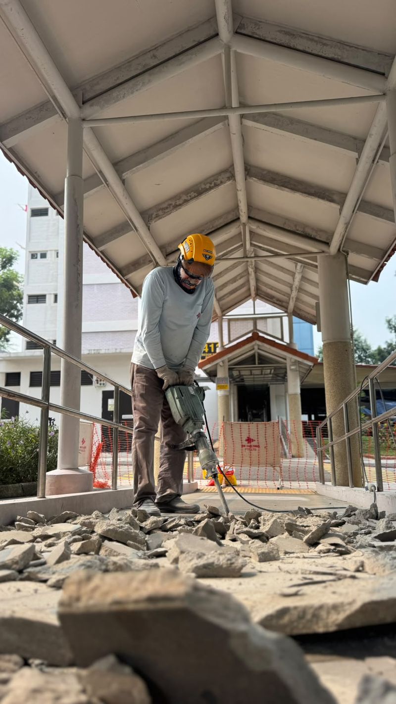 Worker in a yellow helmet using an electric breaker on an existing surface under a covered walkway, with broken concrete pieces in the foreground.