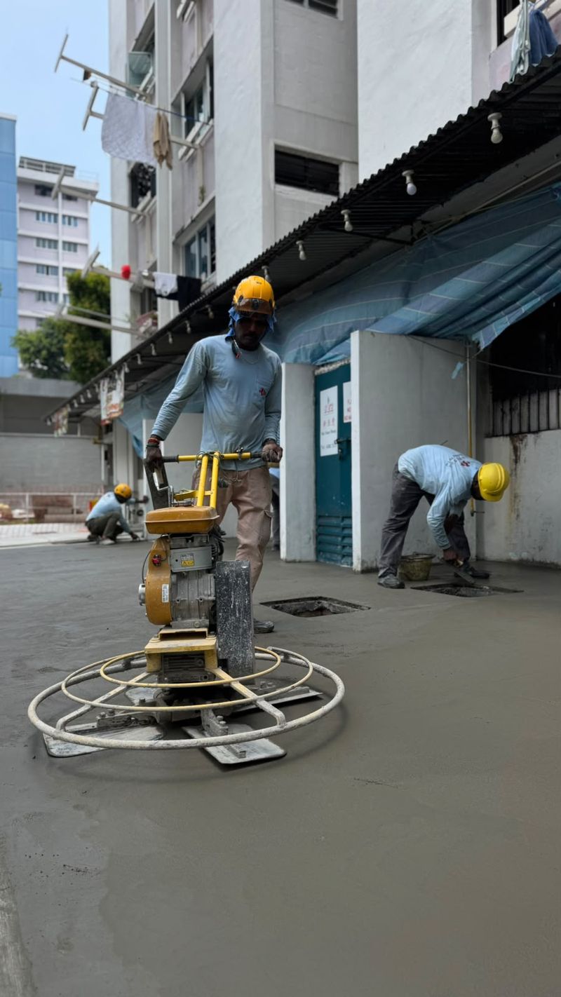 Worker operating a walk-behind power trowel on a freshly placed surface, with a second worker bending over the surface in the background.