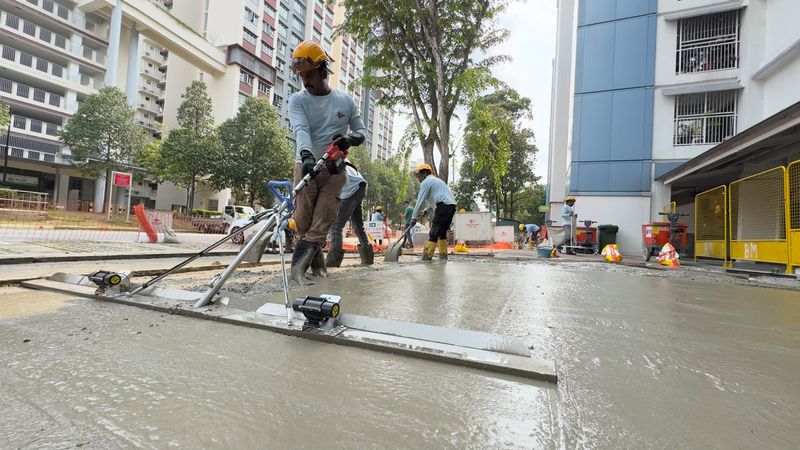 Workers in yellow helmets guiding a powered screed across a freshly placed concrete surface between residential blocks.