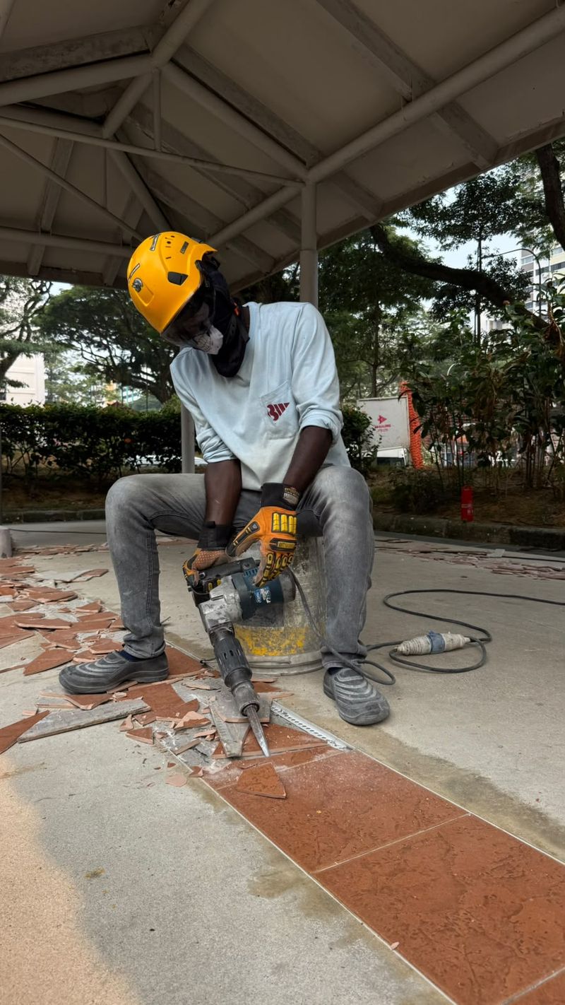 Worker seated on a bucket using a handheld breaker to remove floor tiles, with broken tile pieces around the work area.